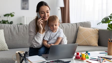 A mother looking at a laptop while being on the phone with a child on her lap. 