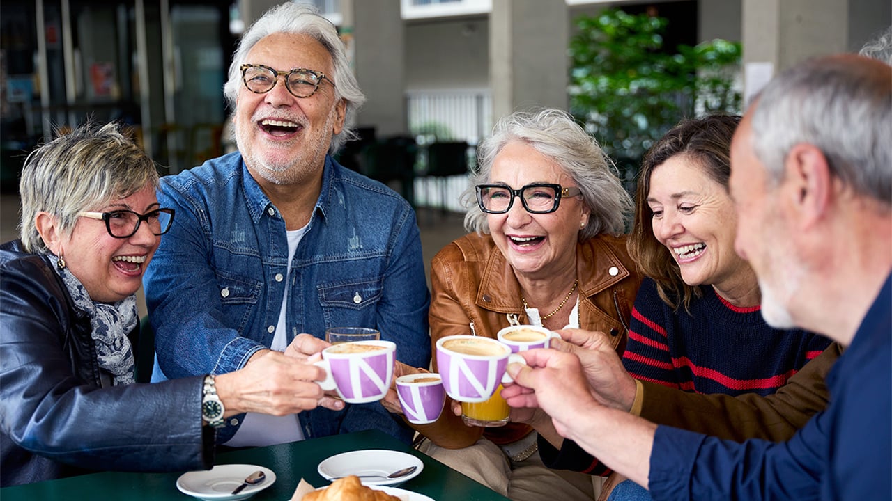 Five people gathered around a table laughing while drinking out of mugs. 