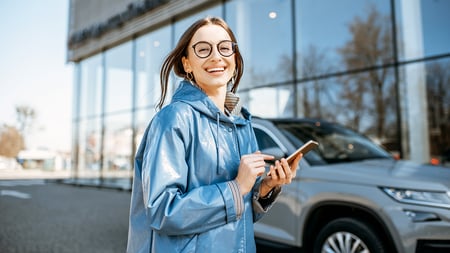 A woman on her phone smiles while standing in front of her new auto