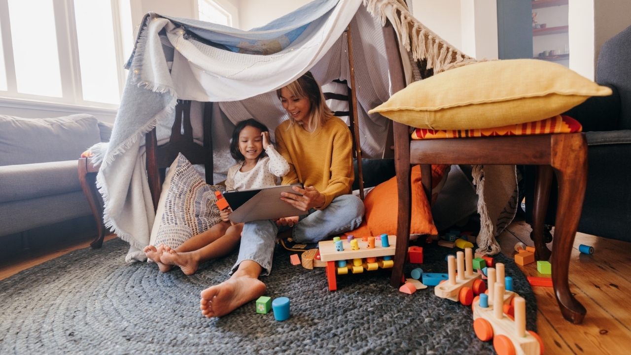 A mother and daughter reading together under a make-shift blanket fort. 